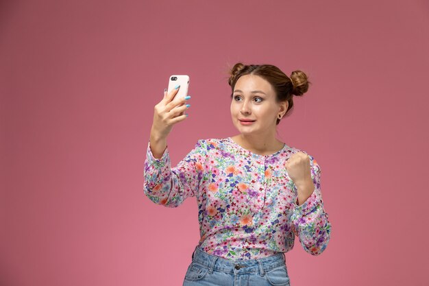 花の正面の若い女性のデザインのシャツとブルージーンズピンクの背景に喜んで式でselfieを取る