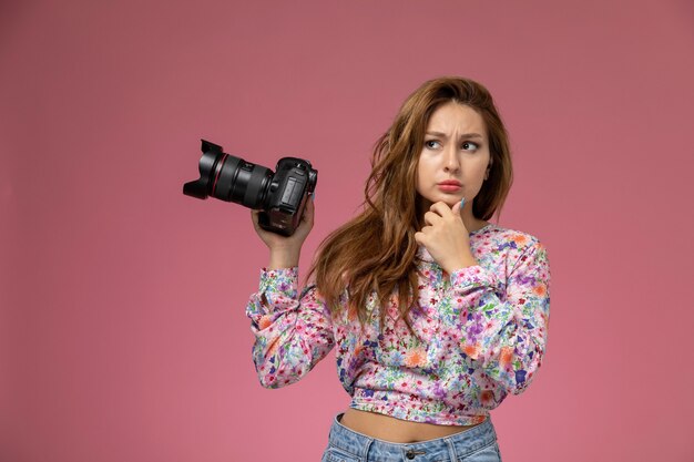 Front view young female in flower designed shirt and blue jeans thinking and holding photo camera on the pink background