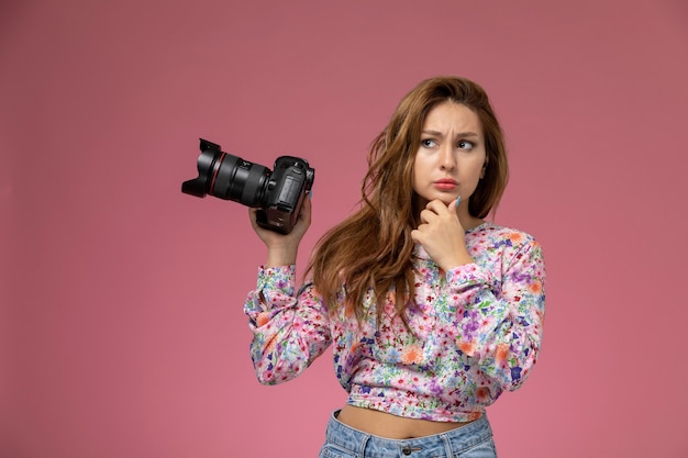 Front view young female in flower designed shirt and blue jeans thinking and holding photo camera on the pink background