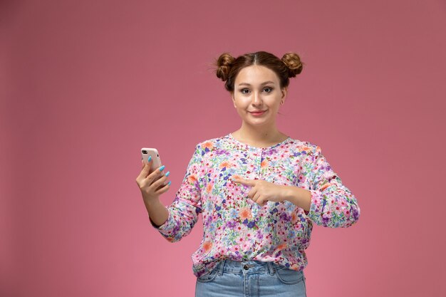 Front view young female in flower designed shirt and blue jeans using a phone smiling on the pink background 