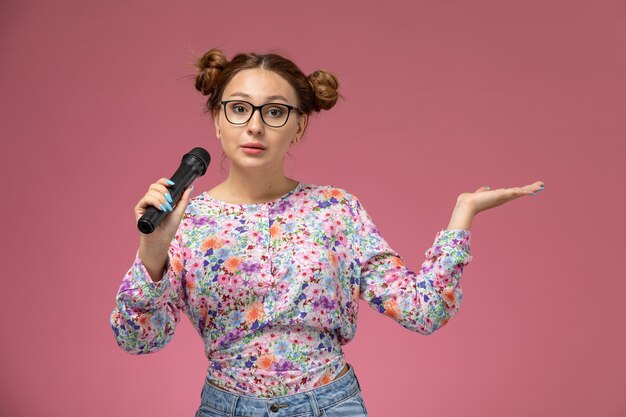 Front view young female in flower designed shirt holding a microphone singing on the pink desk