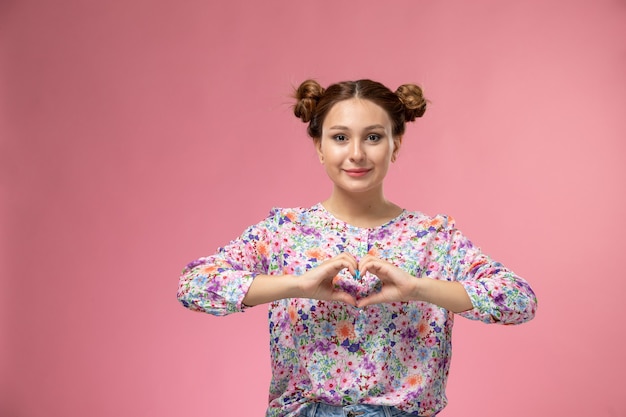 Front view young female in flower designed shirt smiling showing heart sign on the pink background 
