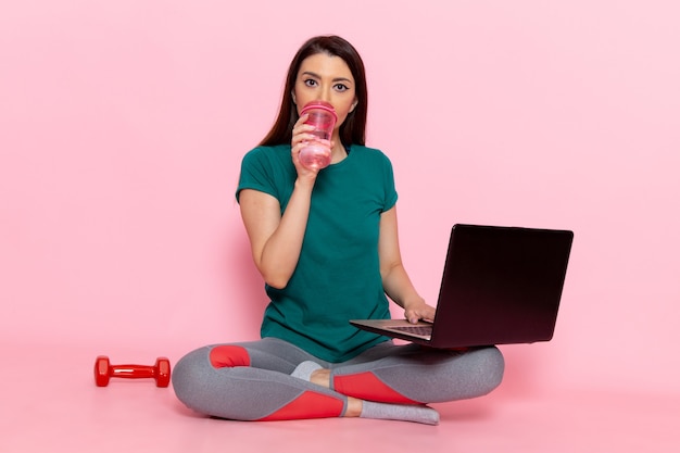 Front view young female in green t-shirt working with her laptop on light pink wall waist sport exercise workouts beauty slim athlete female