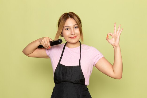Молодая женщина-парикмахер в розовой футболке и черной накидке, вид спереди, поправляет волосы расческой, улыбается на зеленом