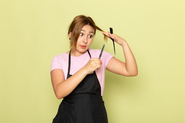Молодая женщина-парикмахер в розовой футболке и черной накидке, держащая кисть и ножницы, фиксирует волосы на зеленом, вид спереди