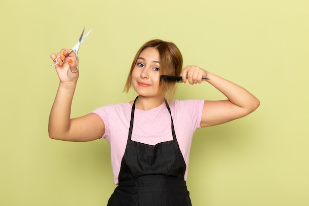 Вид спереди молодой девушки-парикмахера в розовой футболке и черной накидке, держащей кисть и ножницы, поправляющей волосы, улыбаясь на зеленом