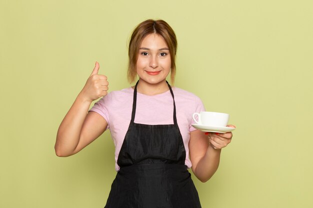 A front view young female hairdresser in pink t-shirt and black cape holding cup and smiling on green