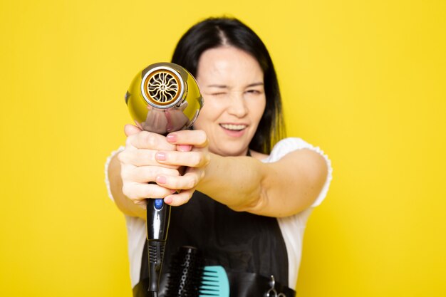 A front view young female hairdresser in white t-shirt black cape holding black hairdryer smiling fun pointing out