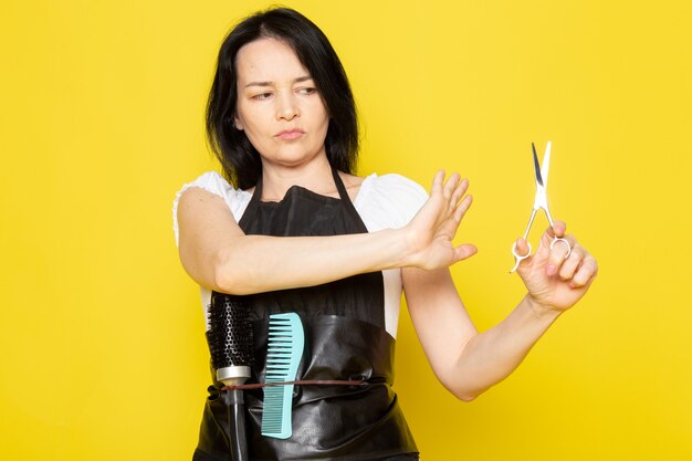 A front view young female hairdresser in white t-shirt black cape holding hair scissors worried expression