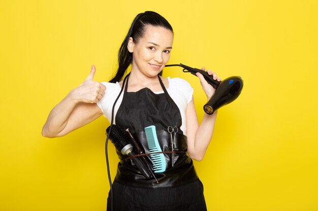 A front view young female hairdresser in white t-shirt black cape holding hairdryer smiling posing