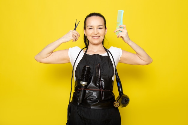 A front view young female hairdresser in white t-shirt black cape holding scissors and brush smiling posing