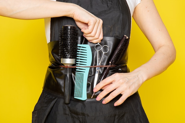 A front view young female hairdresser in white t-shirt black cape taking scissors with brushes wave hair