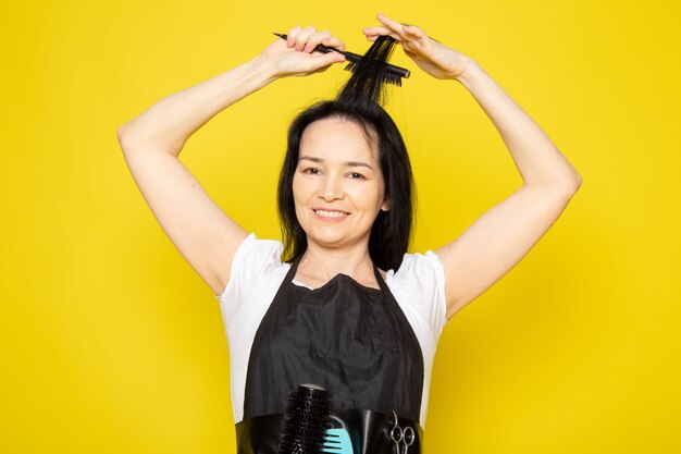 A front view young female hairdresser in white t-shirt black cape using black brush wave hair smiling