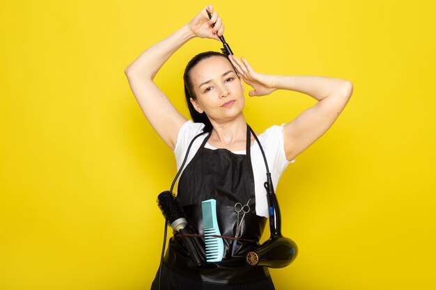 A front view young female hairdresser in white t-shirt black cape with brushes and hairdryer brushing her hair posing