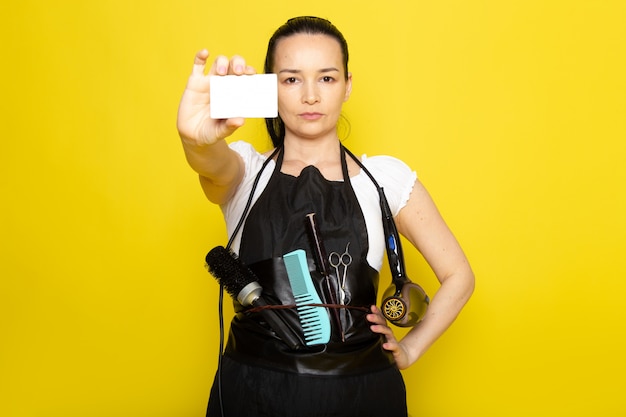 A front view young female hairdresser in white t-shirt black cape with brushes and hairdryer holding white card posing