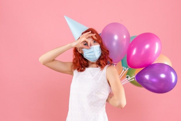 Front view young female hiding colorful balloons in sterile mask on pink desk party covid- new year christmas color