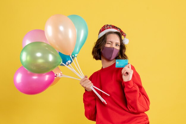 Front view young female holding balloons and bank card in mask on yellow 