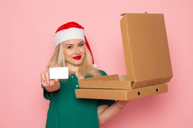 明るいピンクの壁の色の休日のクリスマス新年の写真の仕事の制服に銀行カードとピザボックスを保持している若い女性の正面図