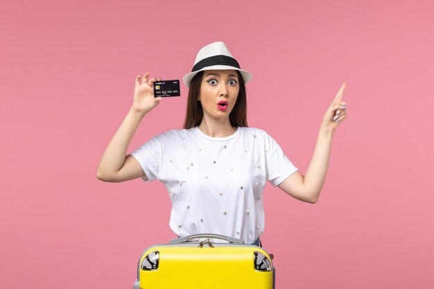 Вид спереди молодая женщина, держащая черную банковскую карту на розовой стене, путешествие летом