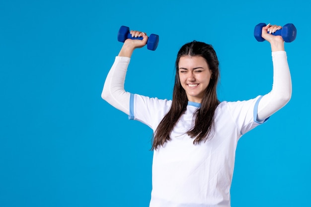 Front view young female holding blue dumbbells on blue wall