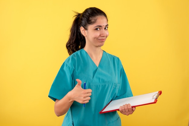 Front view of young female holding clipboard making thumb up on yellow wall