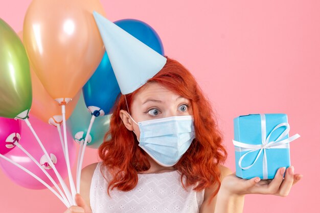 Front view young female holding colorful balloons and little present on pink desk xmas color photo party covid virus