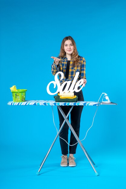 Front view of young female holding the folded clothes sale icon pointing something and standing behind the ironing board in the laundry room