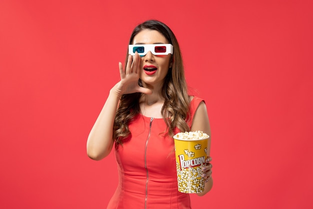 Вид спереди молодая женщина держит попкорн в солнцезащитных очках d на красной поверхности