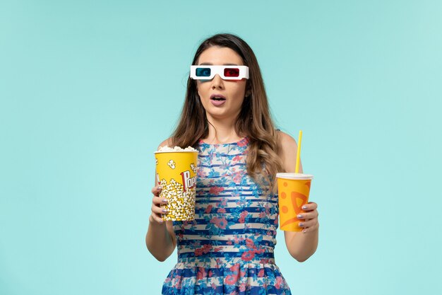 Foto gratuita vista frontale giovane femmina che tiene popcorn e bevanda in occhiali da sole d sulla superficie azzurra