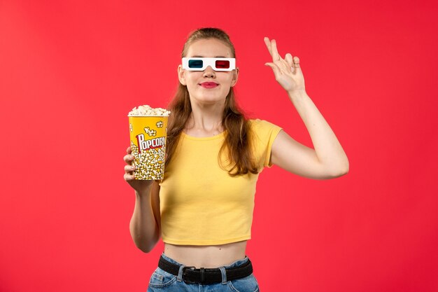 Front view young female holding popcorn package in d sunglasses on light-red wall theater movies cinema film