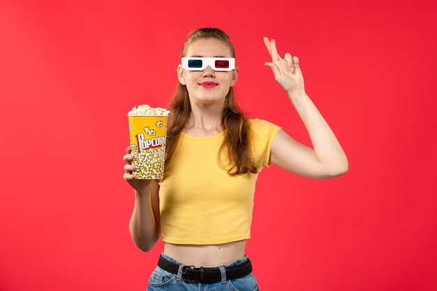 明るい赤の壁の劇場映画シネマフィルムにdサングラスでポップコーンパッケージを保持している正面図若い女性
