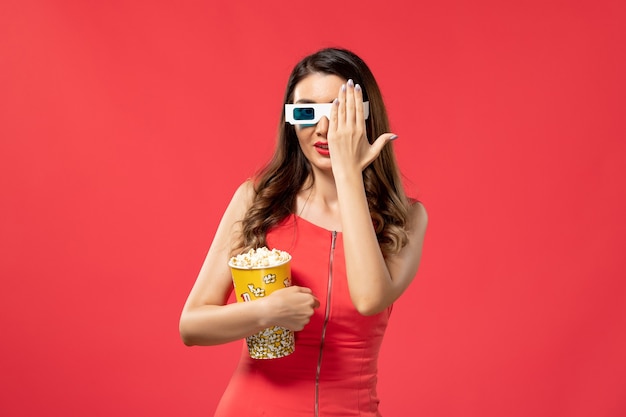 Вид спереди молодая женщина, держащая пакет попкорна в солнцезащитных очках d на красном столе