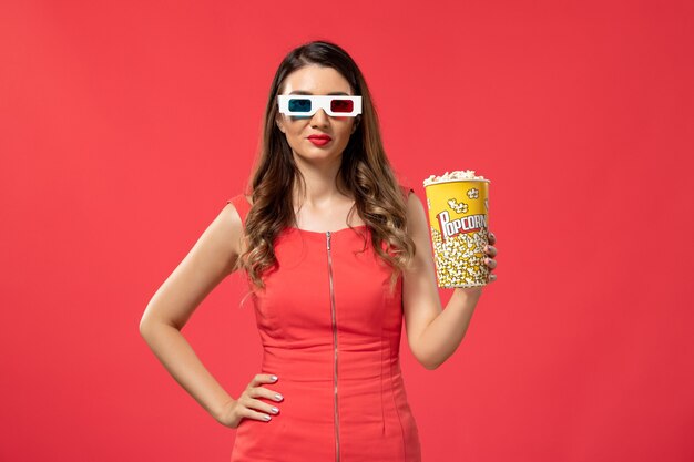 Вид спереди молодая женщина, держащая пакет попкорна в солнцезащитных очках d на красной поверхности