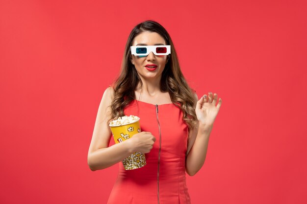 Front view young female holding popcorn package in d sunglasses on the red surface