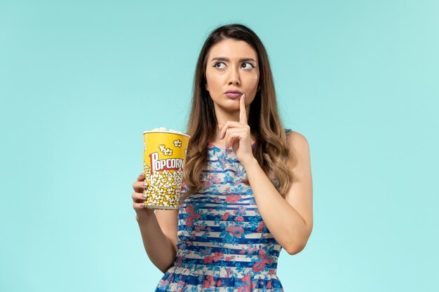 Вид спереди молодая женщина держит пакет попкорна и думает на синей поверхности