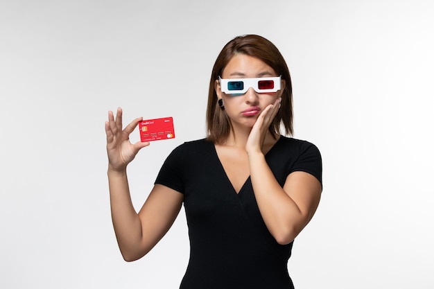Вид спереди молодая женщина держит красную банковскую карту в солнцезащитных очках d на белой поверхности