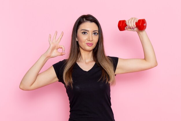 明るいピンクの壁のアスリートスポーツ運動健康トレーニングに赤いダンベルを保持している正面図若い女性