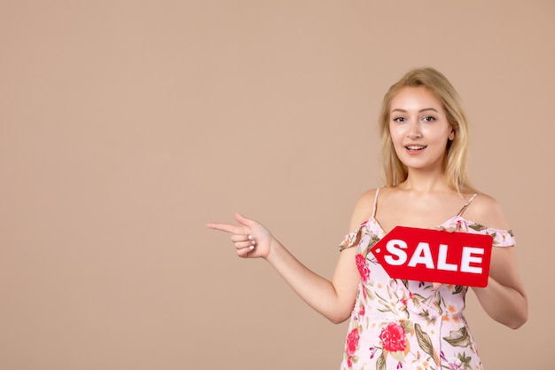 Front view of young female holding red sale board on brown wall