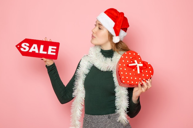 Free photo front view young female holding red sale writing and present on pink wall christmas new year shopping emotion holidays