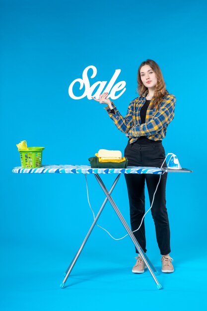 Front view of young female holding the sale icon and standing behind the ironing board in the laundry room