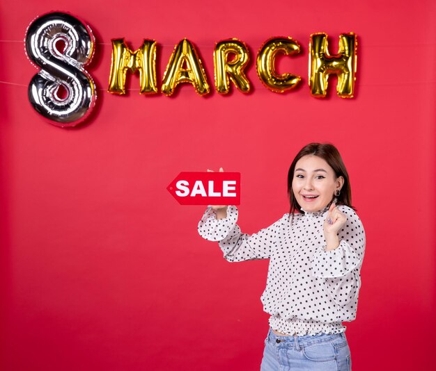 Free photo front view young female holding sale nameplate on march decorated red background shopping love affectionate equality woman holiday feminine beauty