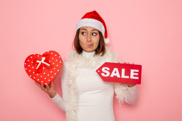 Front view young female holding sale writing and present on pink wall snow christmas color holiday new year fashion