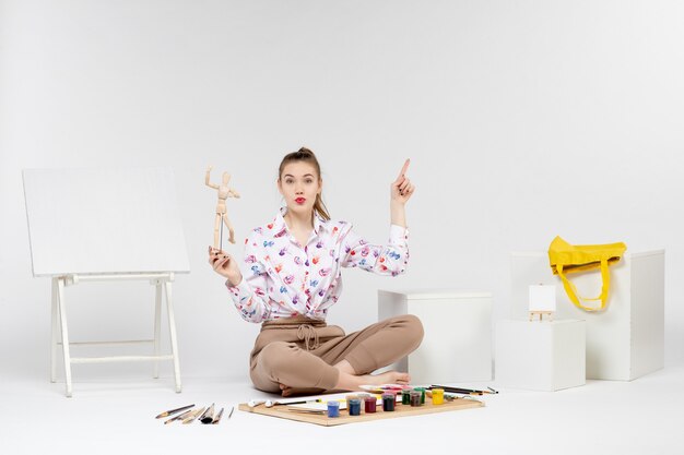 正面図白い机の上のおもちゃの人物像を保持している若い女性アーティストカラーアート女性イーゼル画家描画