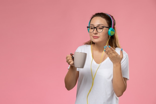 イヤホンを介して音楽を聴くとピンクの背景にお茶とカップを保持している白いtシャツの正面の若い女性