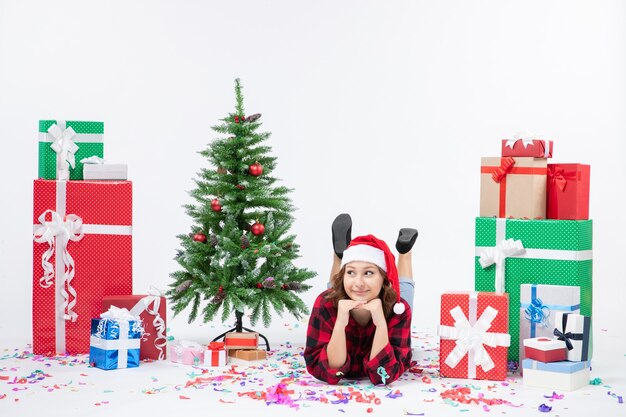 Вид спереди молодая женщина, лежащая вокруг рождественских подарков и праздничной елки на белом фоне, подарок, рождество, новогодние цвета, снег