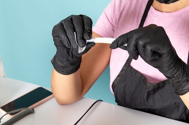 A front view young female manicure in pink t-shirt and black cape with black gloves holding manicure details on blue