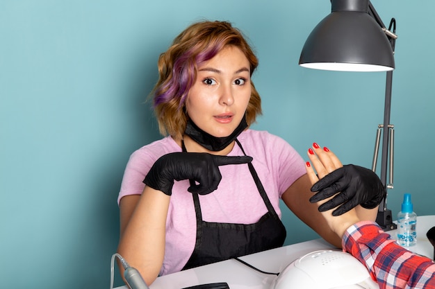 Free photo a front view young female manicure in pink t-shirt with black gloves and black mask doing manicure on blue