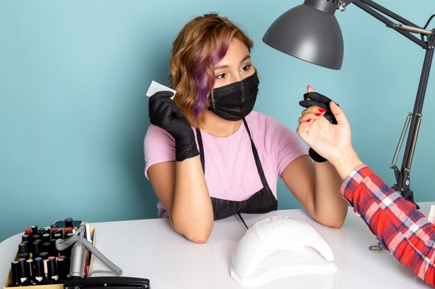 A front view young female manicure in pink t-shirt with black gloves and black mask doing manicure on blue