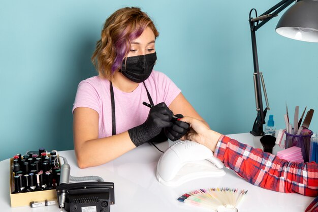 A front view young female manicure in pink t-shirt with black gloves and black mask doing manicure on blue