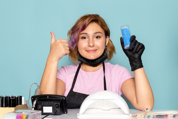 Una giovane donna manicure vista frontale in maglietta rosa con guanti neri e maschera nera in posa e mostrando come segno sul blu